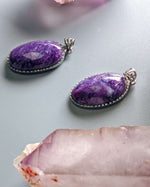 Load image into Gallery viewer, Two purple gemstone pendants on a light gray background with a crystal in the foreground.