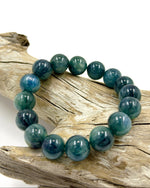 Load image into Gallery viewer, A beaded stretch bracelet made of polished round teal-blue and dark green stones, displayed on a piece of weathered driftwood against a white background.