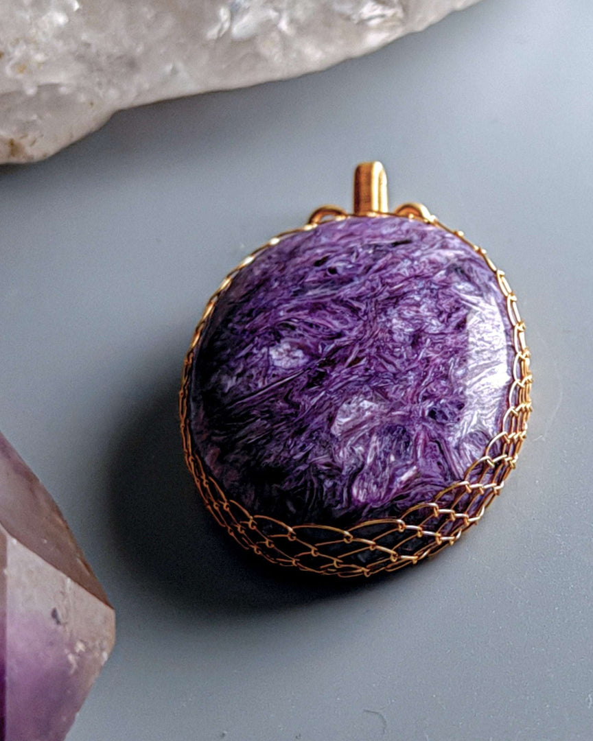 Charoite Pendant (Oval Wire-Wrapped) on a gray surface with crystals.