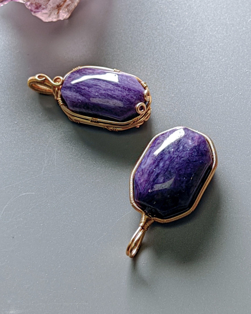 Two purple stone pendants with gold wire wrapping on a gray surface