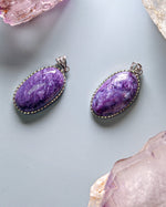 Load image into Gallery viewer, Two purple stone pendants on a light gray surface with crystals around.
