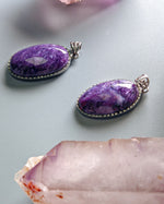 Load image into Gallery viewer, Two purple gemstone pendants on a light gray background with a crystal in the foreground.
