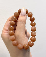 A beaded stretch bracelet made of smooth, polished round beads in a warm peachy-brown color, held up by a hand against a plain light gray background.