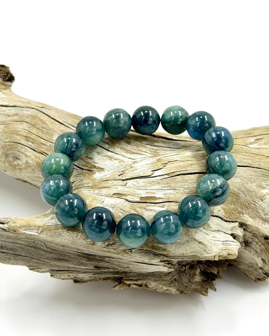 A beaded stretch bracelet made of polished round teal and blue-green stones, displayed on a piece of weathered driftwood against a clean white background.