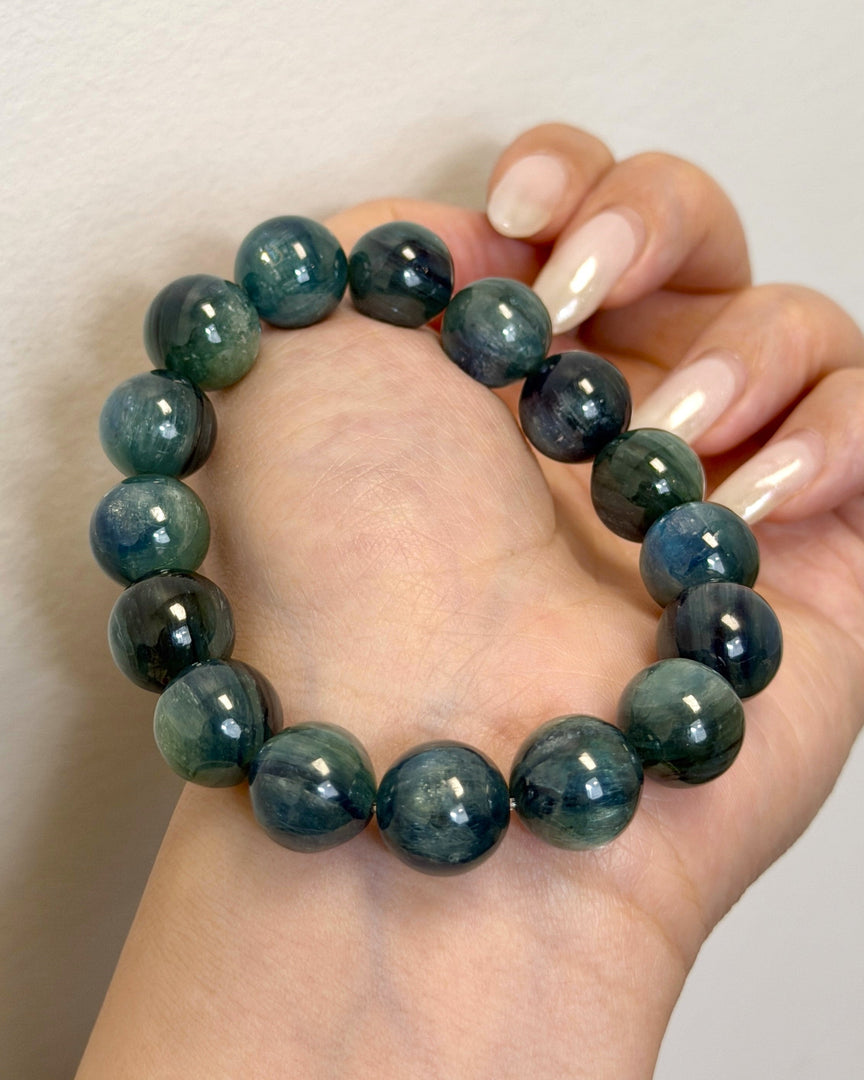 A beaded bracelet made of polished dark teal and green round stones, worn on a person's wrist with manicured nails visible against a plain light background.