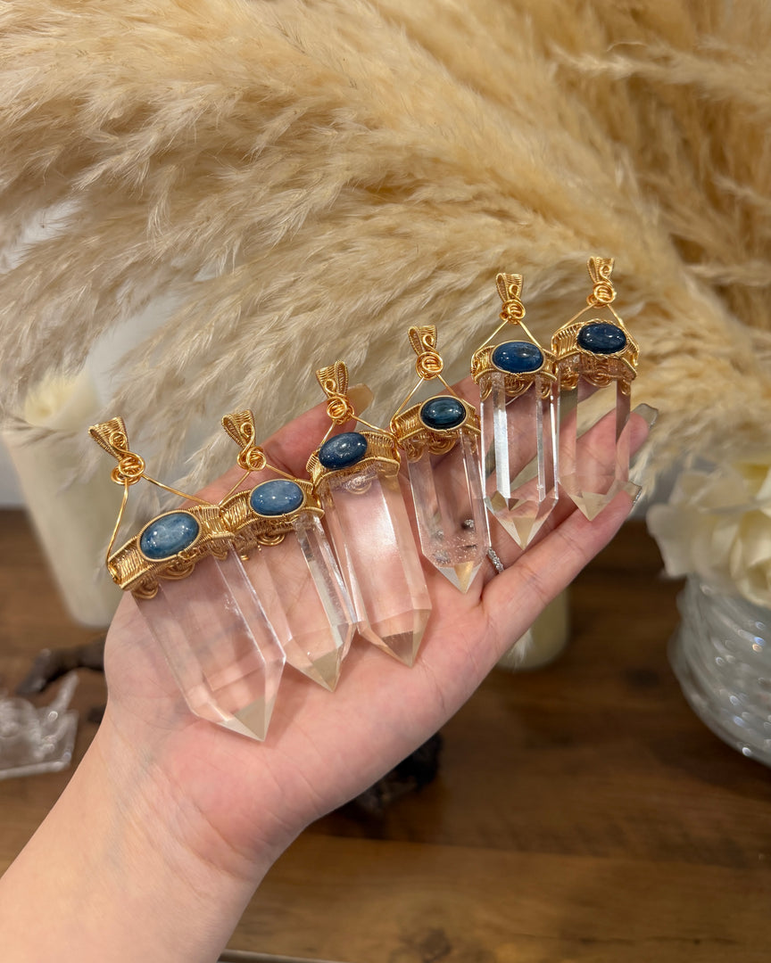 A hand holds five clear crystal point pendants with gold-toned metal caps and oval blue stones, set against a soft beige faux-fur backdrop and wooden surface.